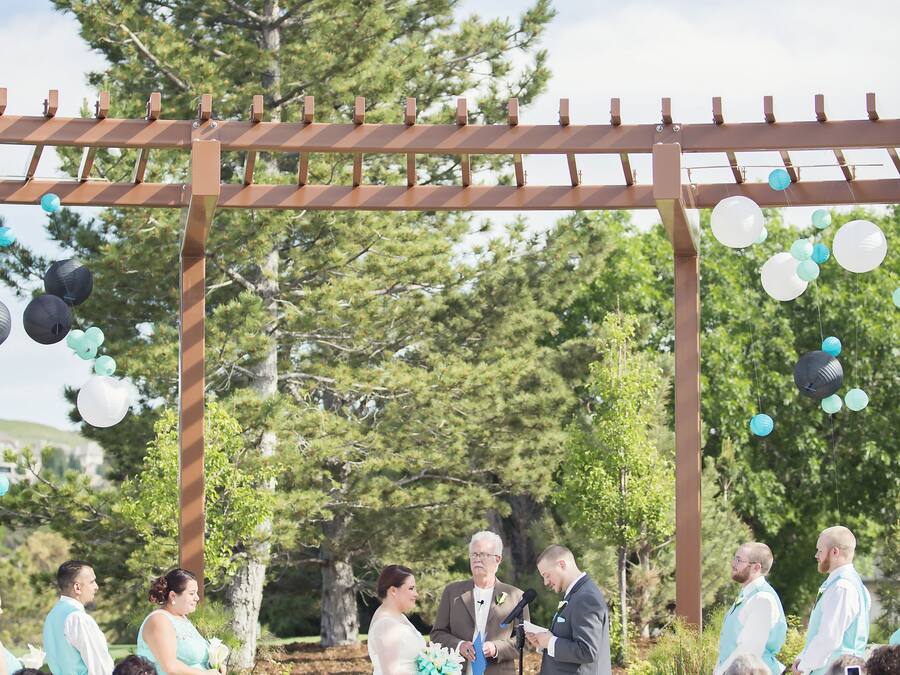 A wedding ceremony outdoors with a wooden pergola and guests seated on white chairs.