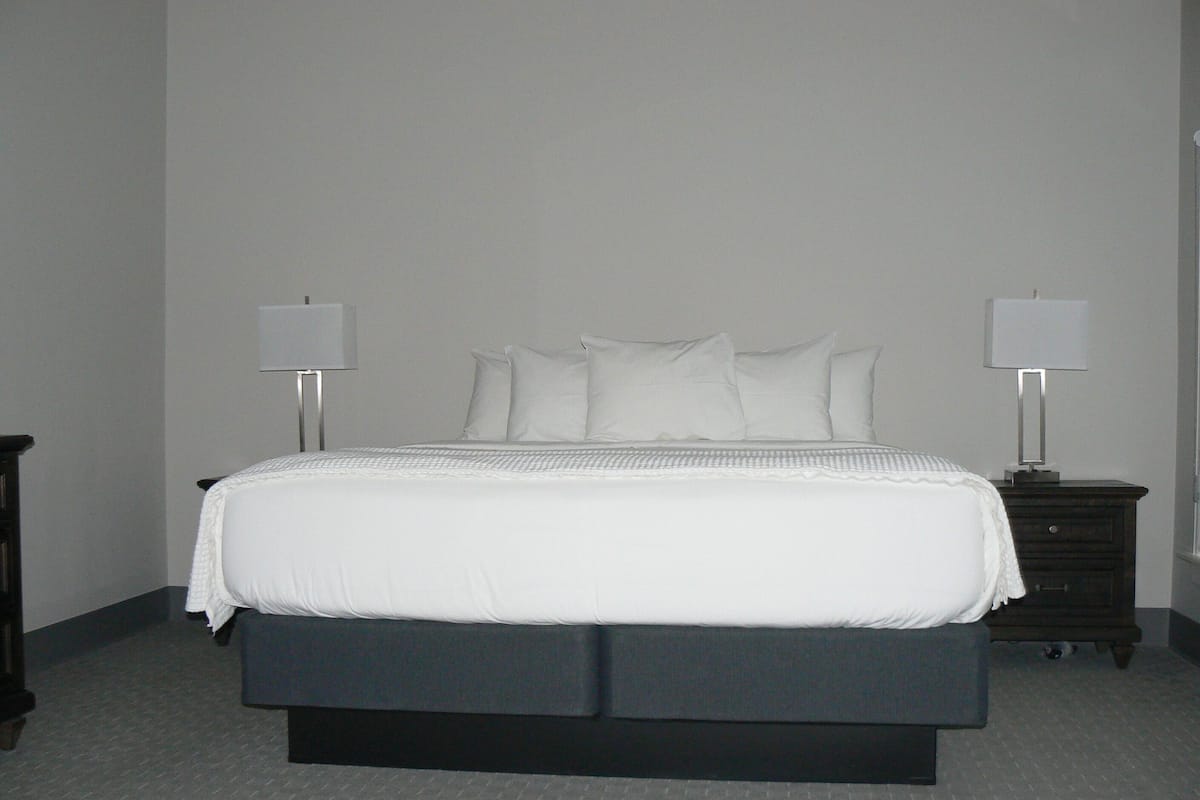 Un lit recouvert d’une couette blanche et agrémenté d’oreillers, avec deux tables de chevet dotées de lampes de chaque côté, dans une pièce aux murs unis et au sol recouvert de moquette.