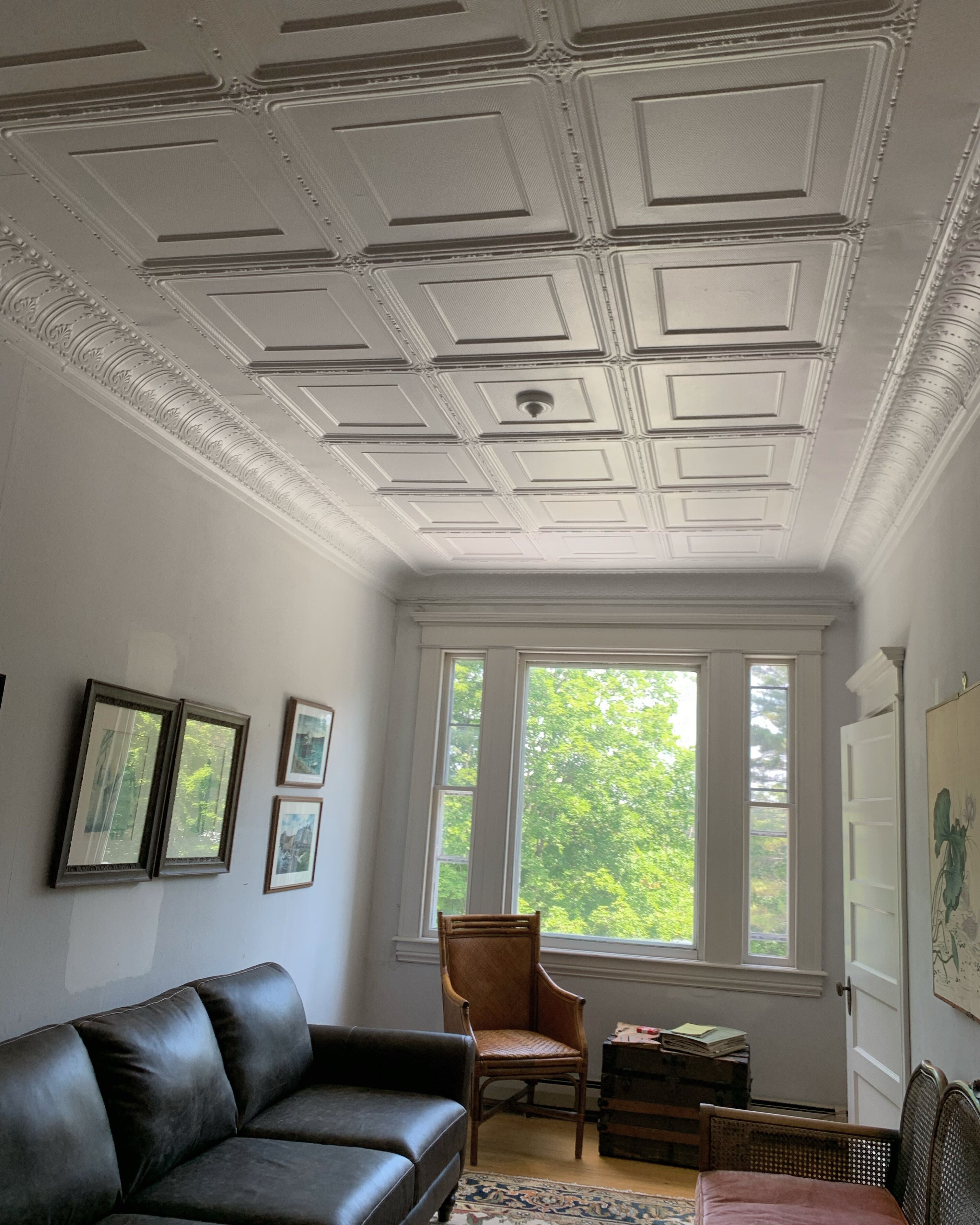 A room with a white ceiling, a brown leather sofa, a wooden chair, a window with a view of trees, and a door with a painting.