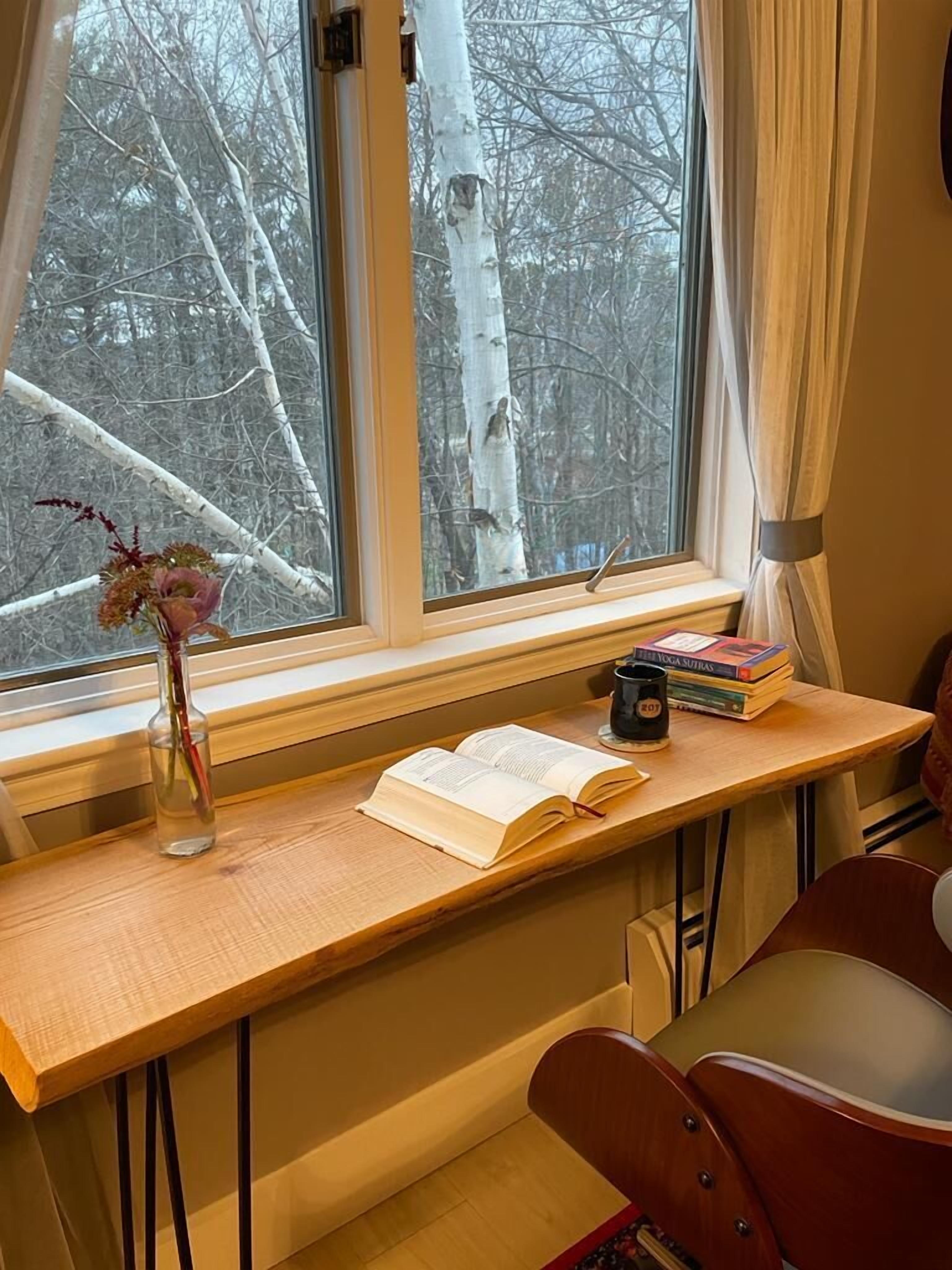 A wooden desk with a book, a mug, and a vase with flowers by a window with a view of trees.