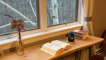 A wooden desk with a book, a mug, and a vase with flowers by a window with a view of trees.