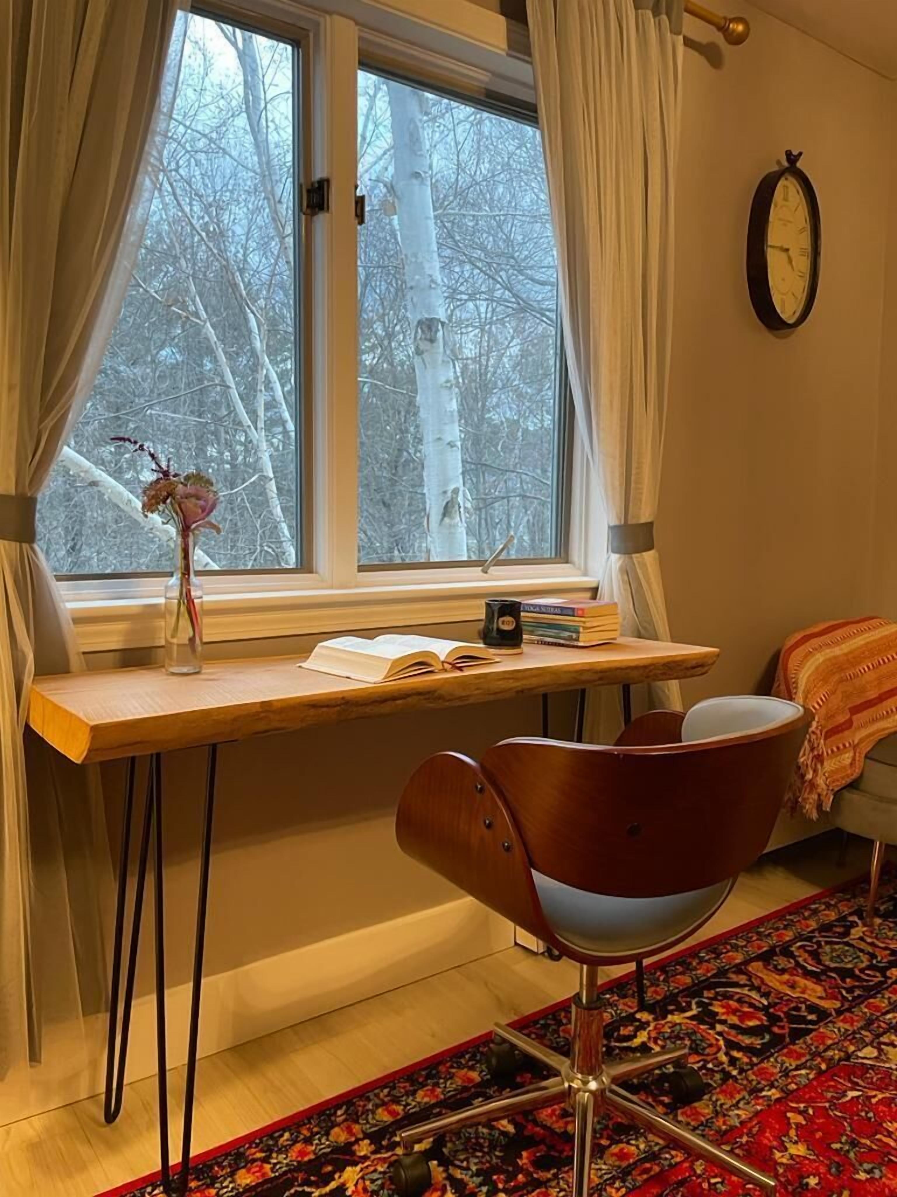 Ett mysigt rum med ett skrivbord i trä, en stol, ett fönster med gardiner, en klocka och en mönstrad matta.
