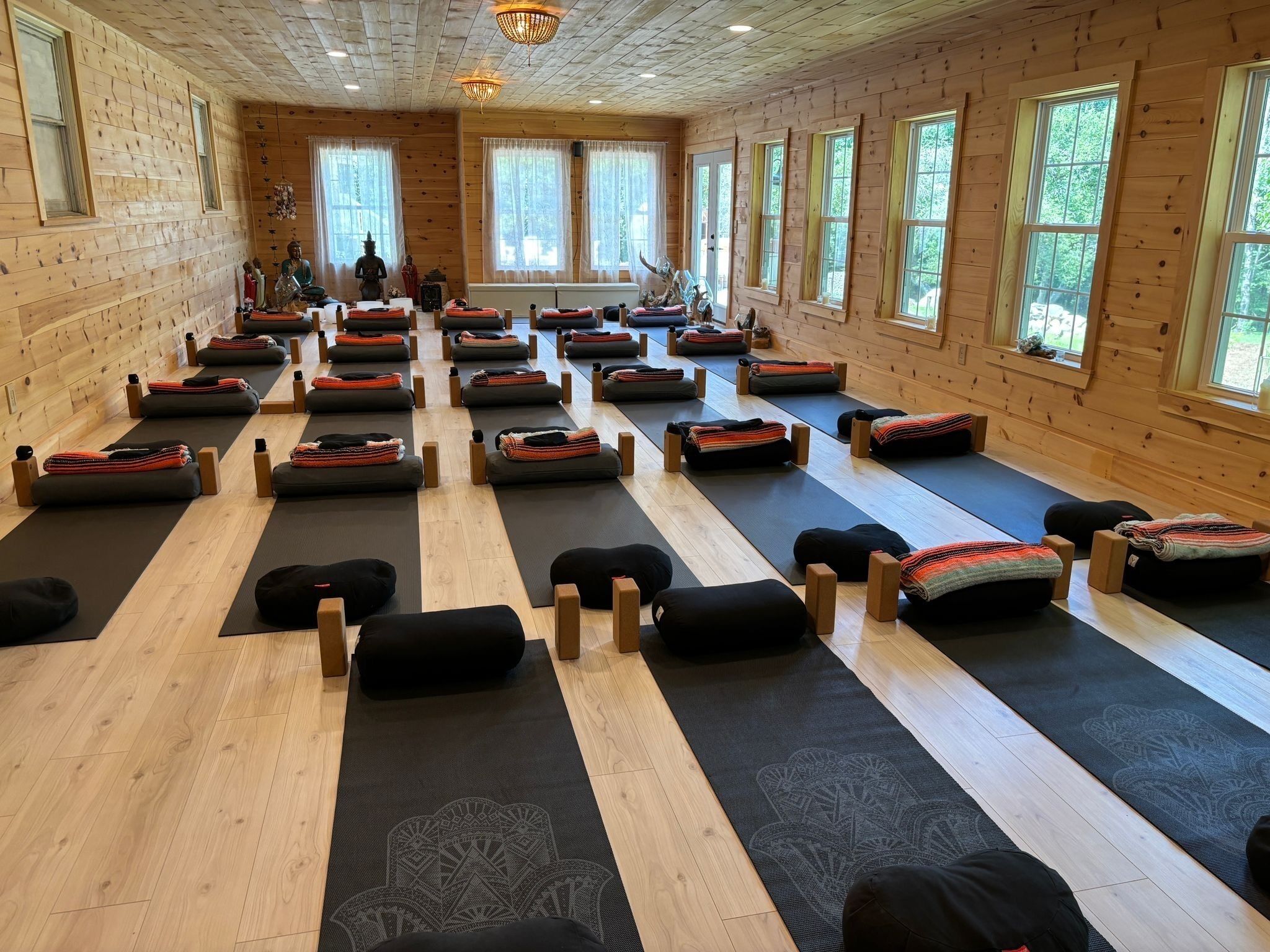 A yoga studio with multiple mats and cushions arranged in rows, wooden walls, and large windows.