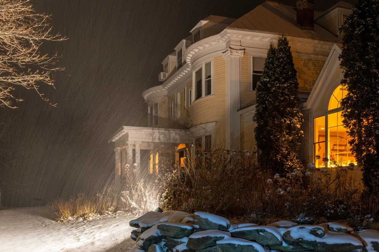 Una casa grande y ornamentada con un patio delantero cubierto de nieve y una ventana iluminada, lo que sugiere un ambiente invernal.