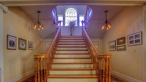 Uma escadaria imponente com balaústres de madeira e corrimão, que leva a um aposento com uma grande janela em arco e um candelabro.