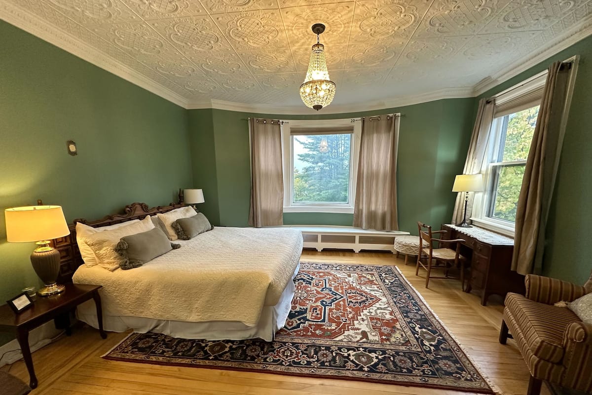 Un dormitorio con una cama grande, un escritorio, una silla, un ventanal con cortinas, un candelabro y una alfombra estampada.
