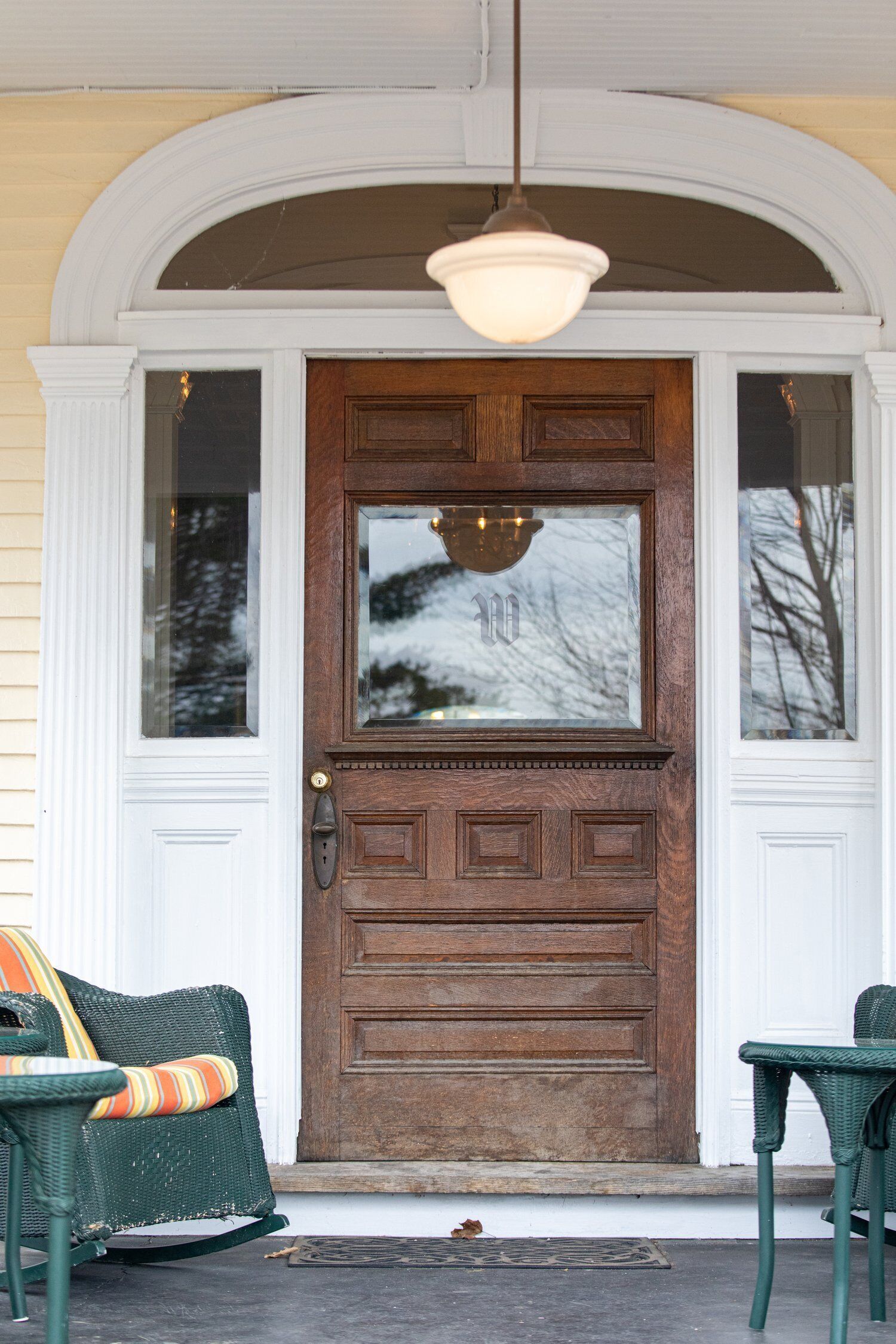 A wooden front door with a glass panel, flanked by white doors, and two wicker chairs with cushions in front.