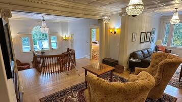 A spacious living room with a high ceiling, ornate moldings, and a large chandelier. There is a wooden coffee table, a patterned sofa, and a dining area with a wooden table and chairs.
