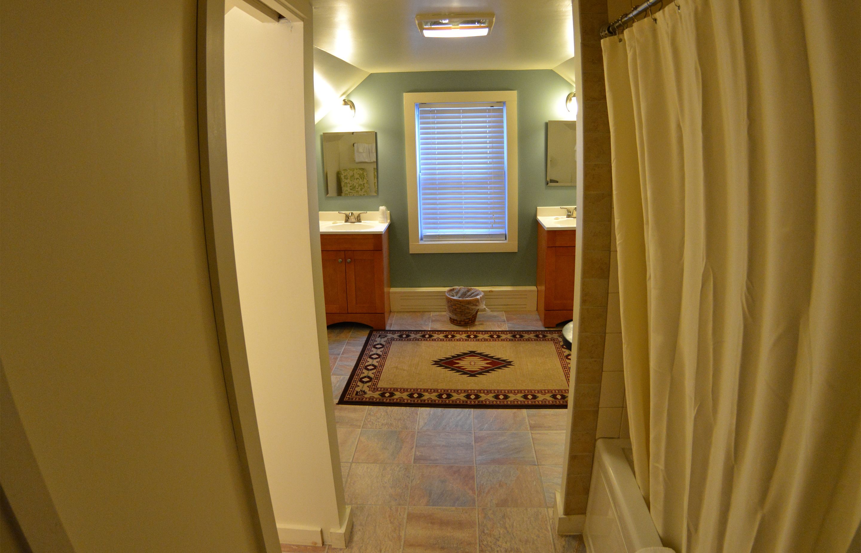 A bathroom with two sinks, a window with blinds, a rug, and a shower curtain.