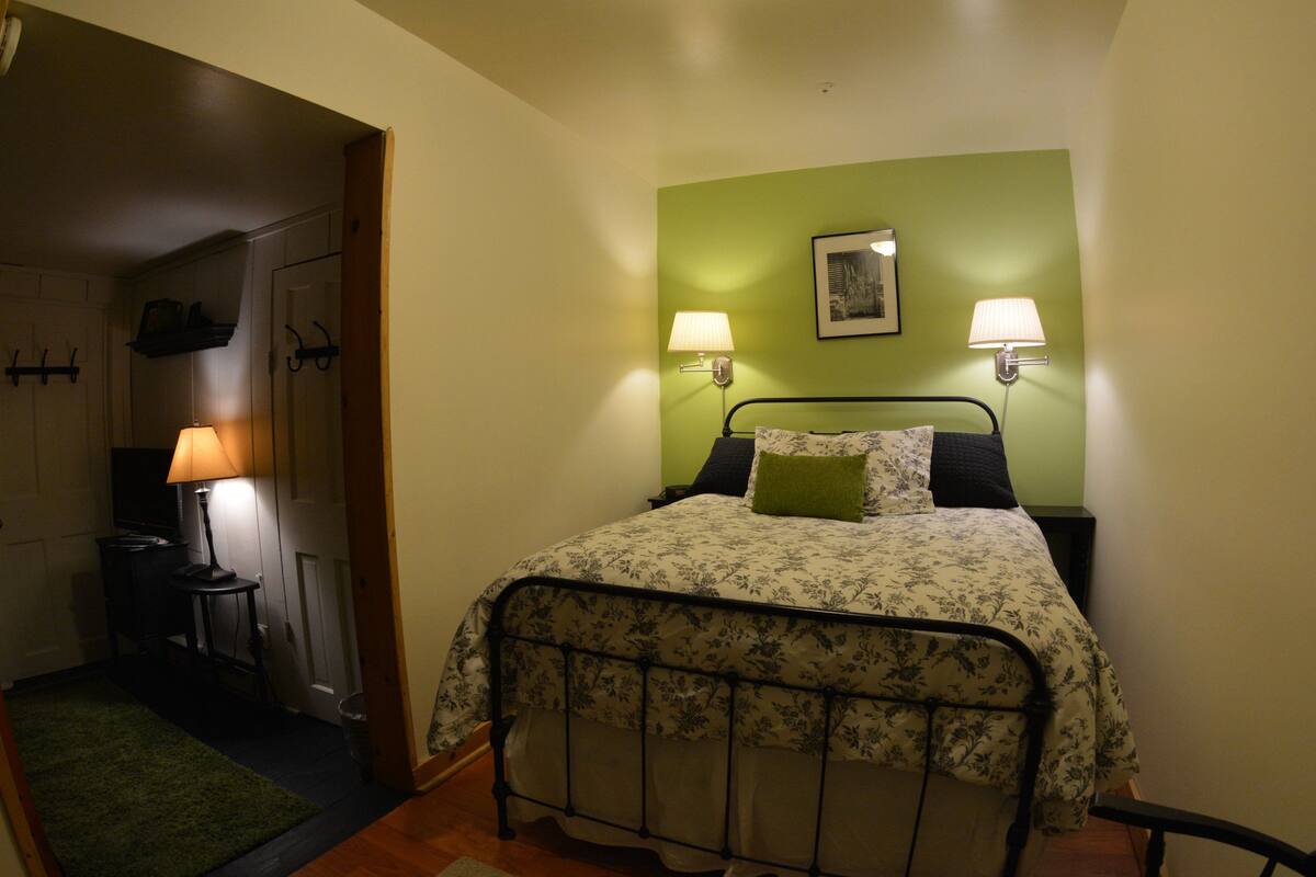 Quarto com cama, duas luminárias de cabeceira, parede verde e porta que dá para outro cômodo.
