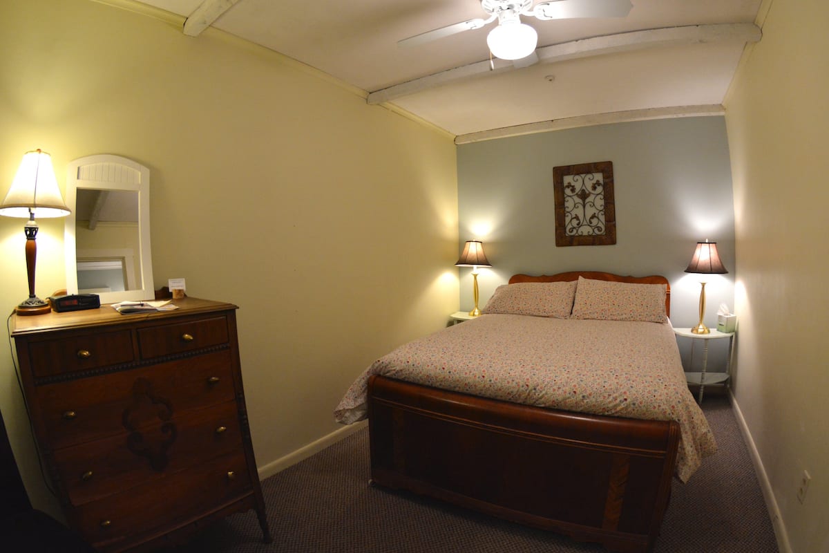 Quarto com cama de madeira, cómoda, duas luminárias de apoio e ventoinha no teto.