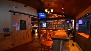 A bar area with wooden flooring, multiple bar stools, and a counter. There are two televisions mounted on the wall, and a sign indicating a 5% discount on Wednesdays.