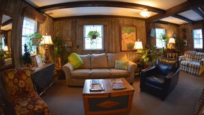 A cozy living room with a sofa, armchair, and coffee table, wooden walls, and large windows with plants.