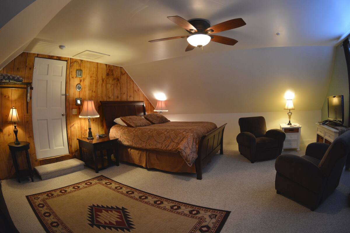 Um quarto com uma cama, duas cadeiras, uma escrivaninha, um ventilador de teto e duas luminárias de cabeceira.