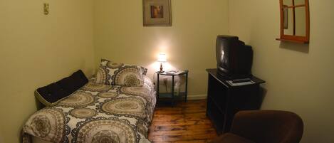 A small, dimly lit room with a bed, a chair, a television, and a small table with a lamp.
