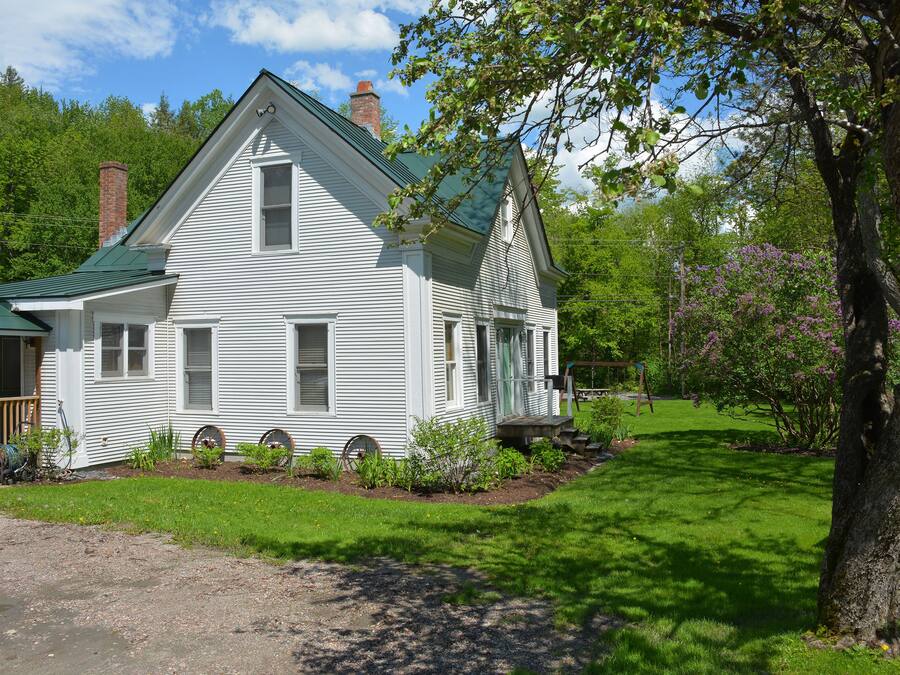 A white two-story house with a green roof, surrounded by a well-maintained lawn and trees.