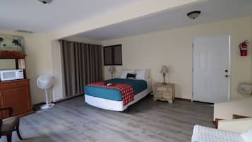 Slaapkamer met een bed, nachtkastjes, een bureau, een stoel, een ventilator, een magnetron, een brandblusser en een deur.