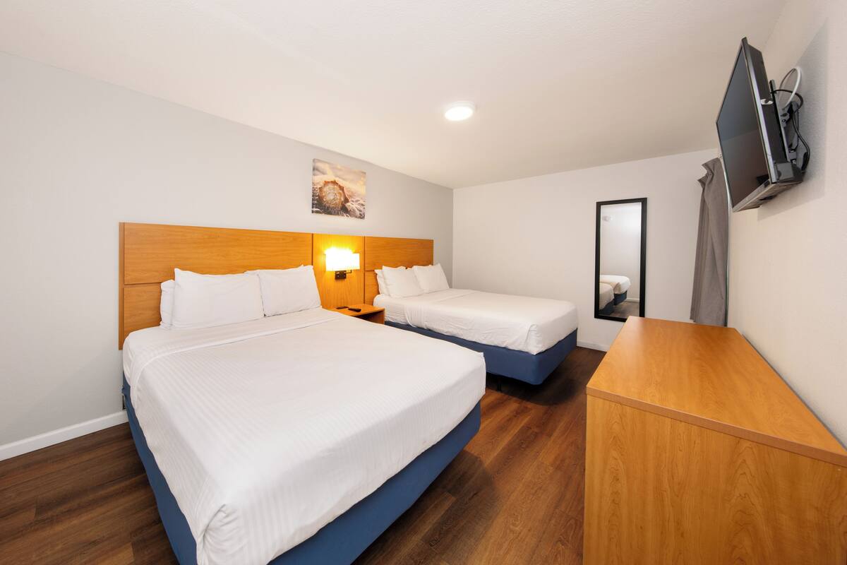 A hotel room with two beds, a wooden headboard, a bedside table with a lamp, a flat-screen TV mounted on the wall, and a mirror.