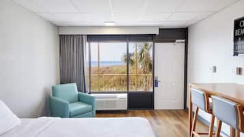 A hotel room with a bed, a chair, a desk, and a large window with a view of the beach.