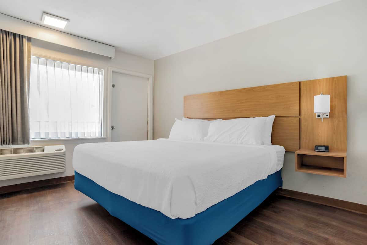 A hotel room with a large bed, a bedside table with a lamp, a window with curtains, and a wall-mounted heater.