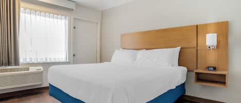 A hotel room with a large bed, a bedside table with a lamp, a window with curtains, and a wall-mounted heater.