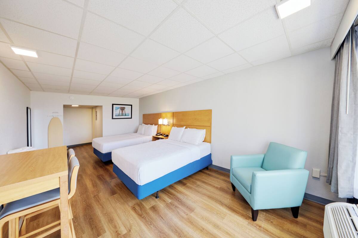 A hotel room with two beds, a wooden desk, a blue armchair, and a desk lamp.