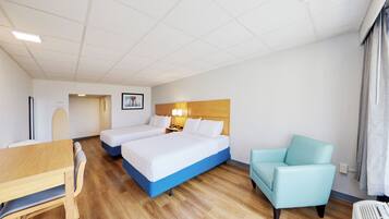 A hotel room with two beds, a wooden desk, a blue armchair, and a desk lamp.