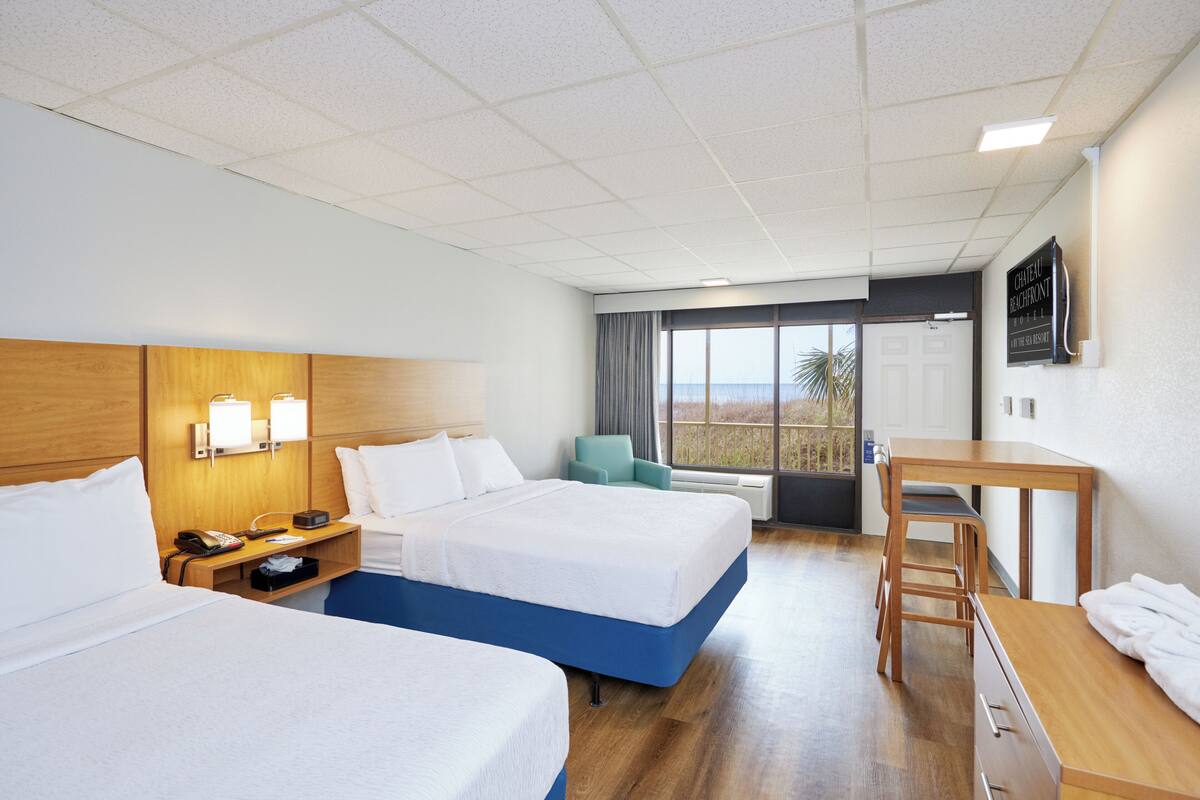 A hotel room with two beds, a desk, a chair, and a view of the outdoors.