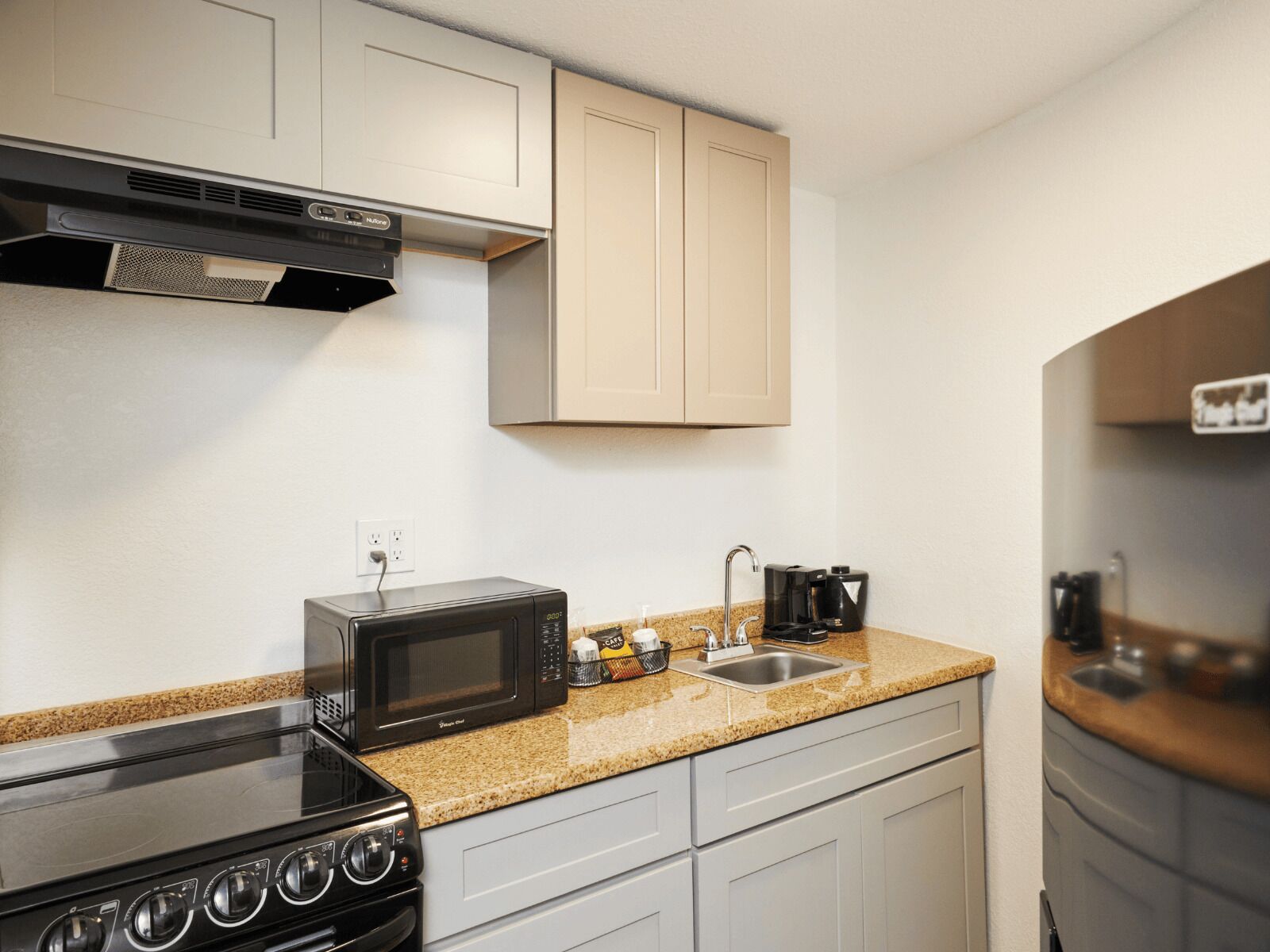 A compact kitchen with a stove, microwave, and sink, featuring light-colored cabinets and a granite countertop.