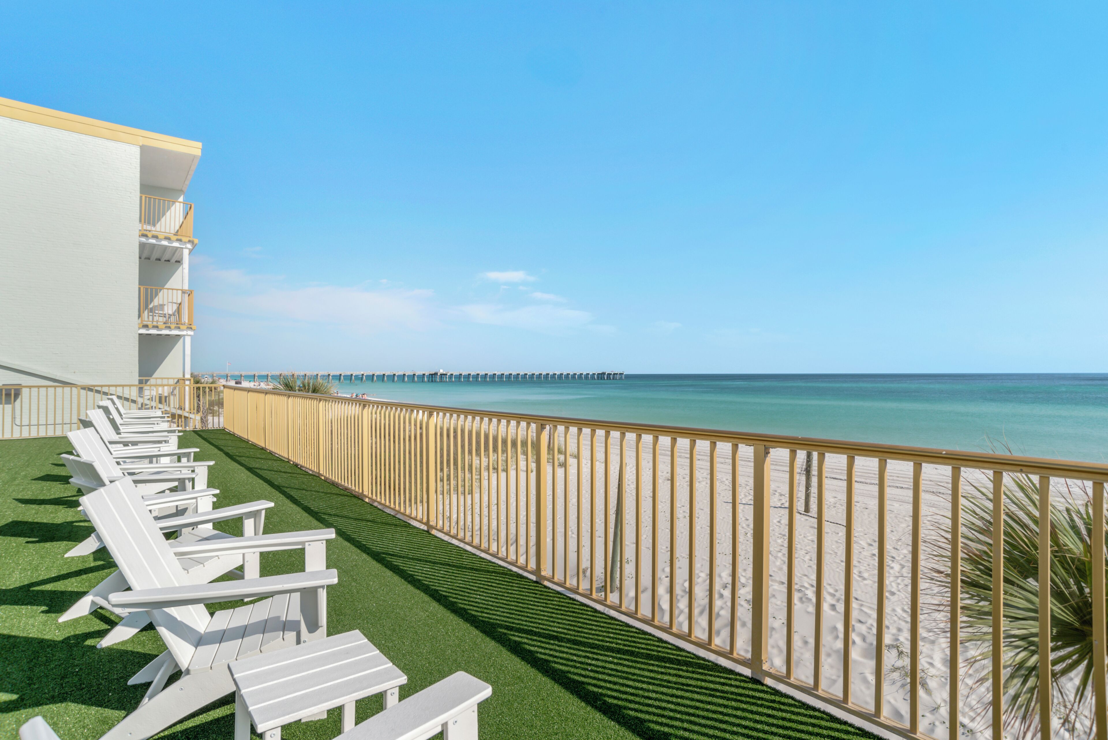 A balcony with white chairs, a view of the beach, and a pier in the distance.