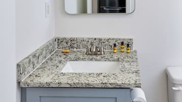 A bathroom with a granite countertop sink, a mirror, a toilet, and a trash bin.