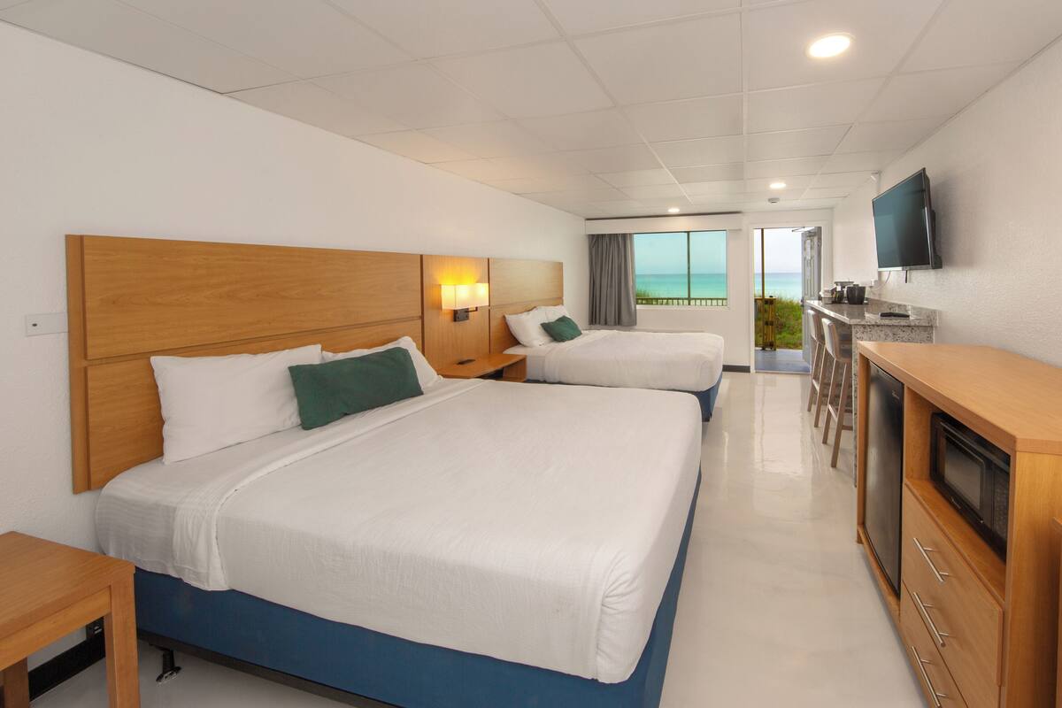 A hotel room with two beds, a TV, a kitchenette, and a view of the ocean.