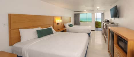 A hotel room with two beds, a TV, a kitchenette, and a view of the ocean.