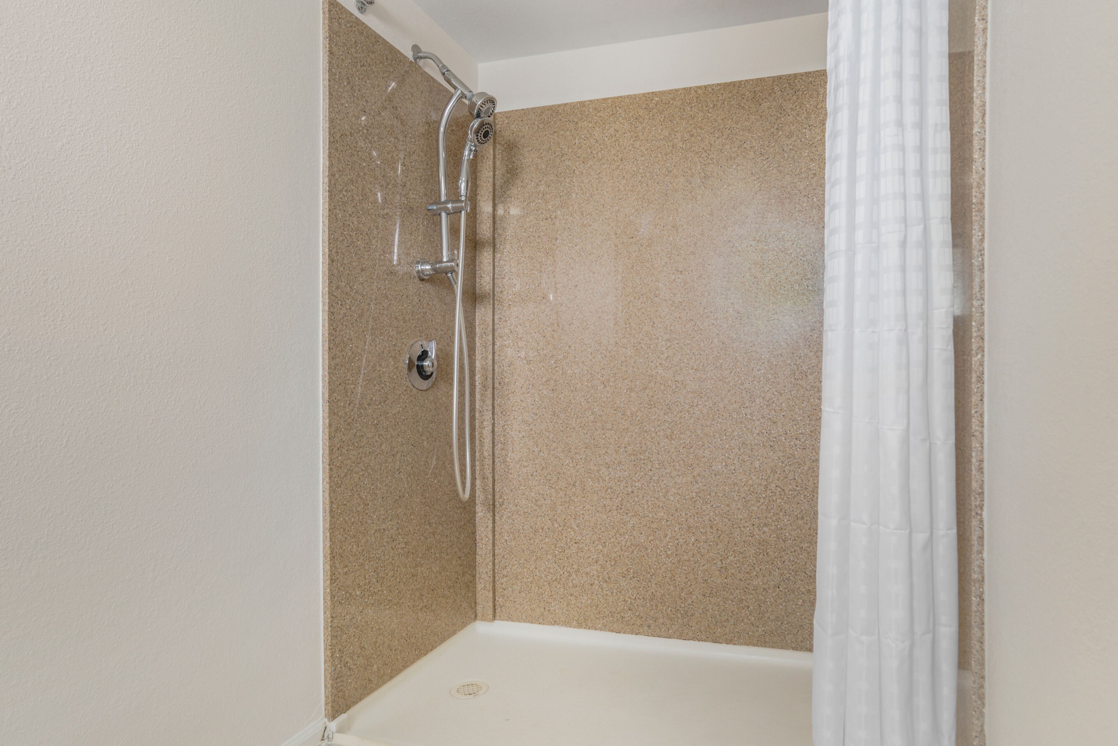 A shower with a handheld showerhead, a wall-mounted faucet, and a white shower curtain.
