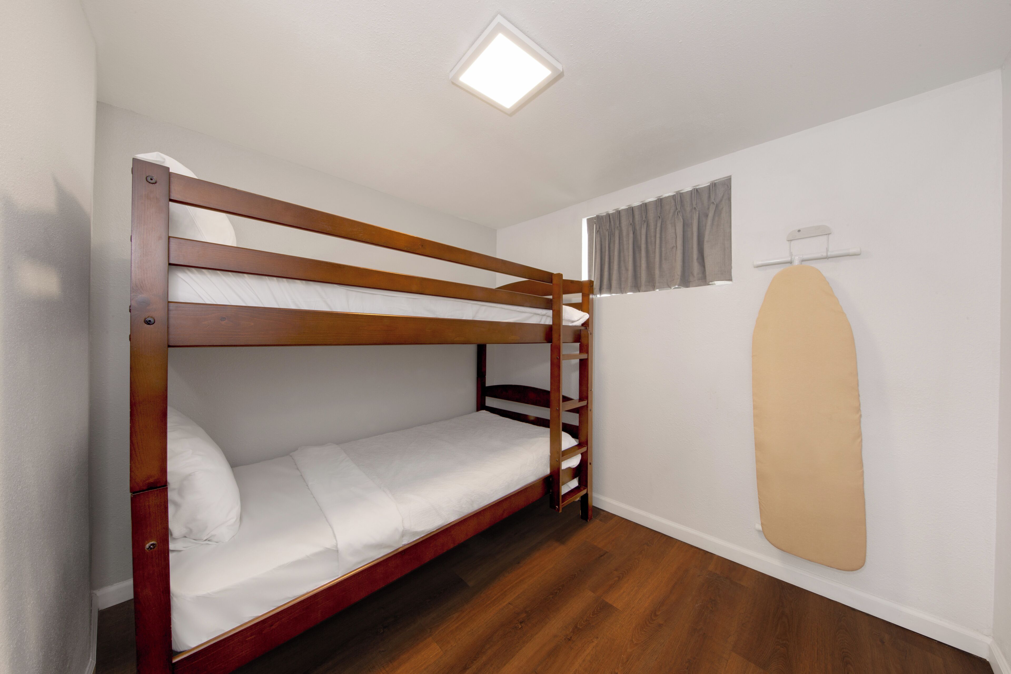 Un lit superposé dans une chambre avec un luminaire au plafond, une fenêtre avec des rideaux et une planche à repasser fixée au mur.