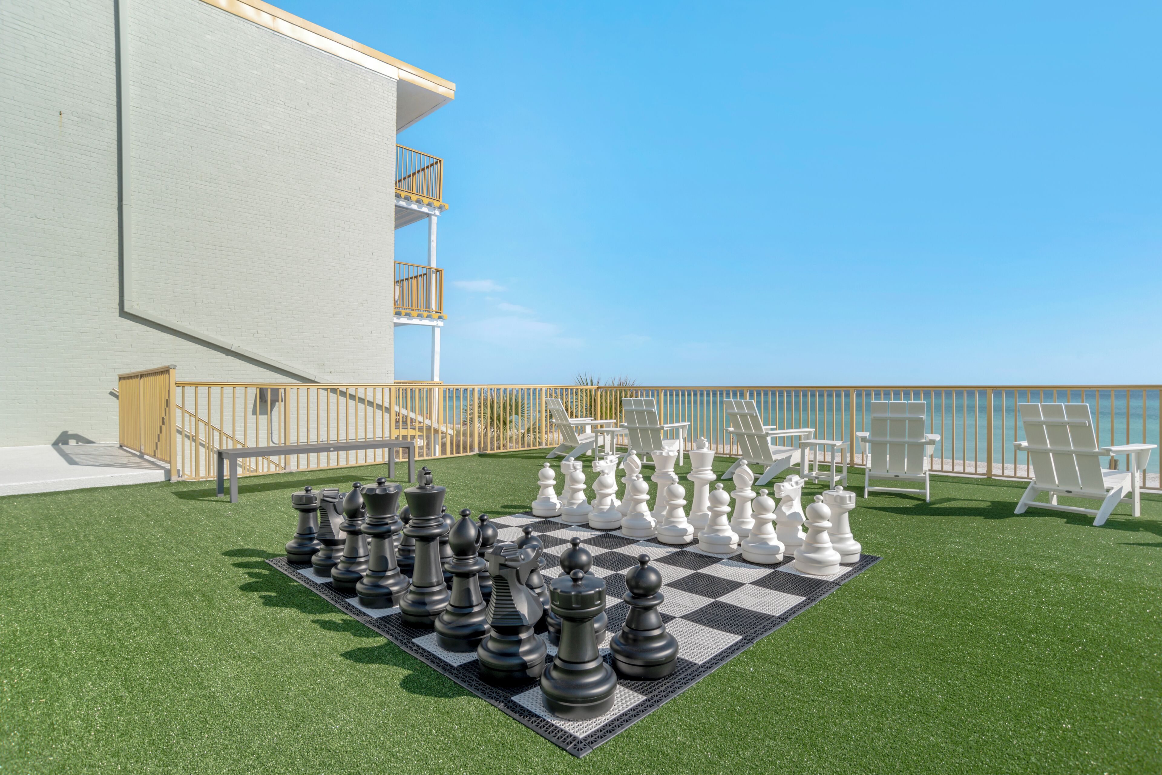 Tablero de ajedrez sobre césped artificial, con piezas blancas y negras, sillas exteriores y un edificio con balcón.