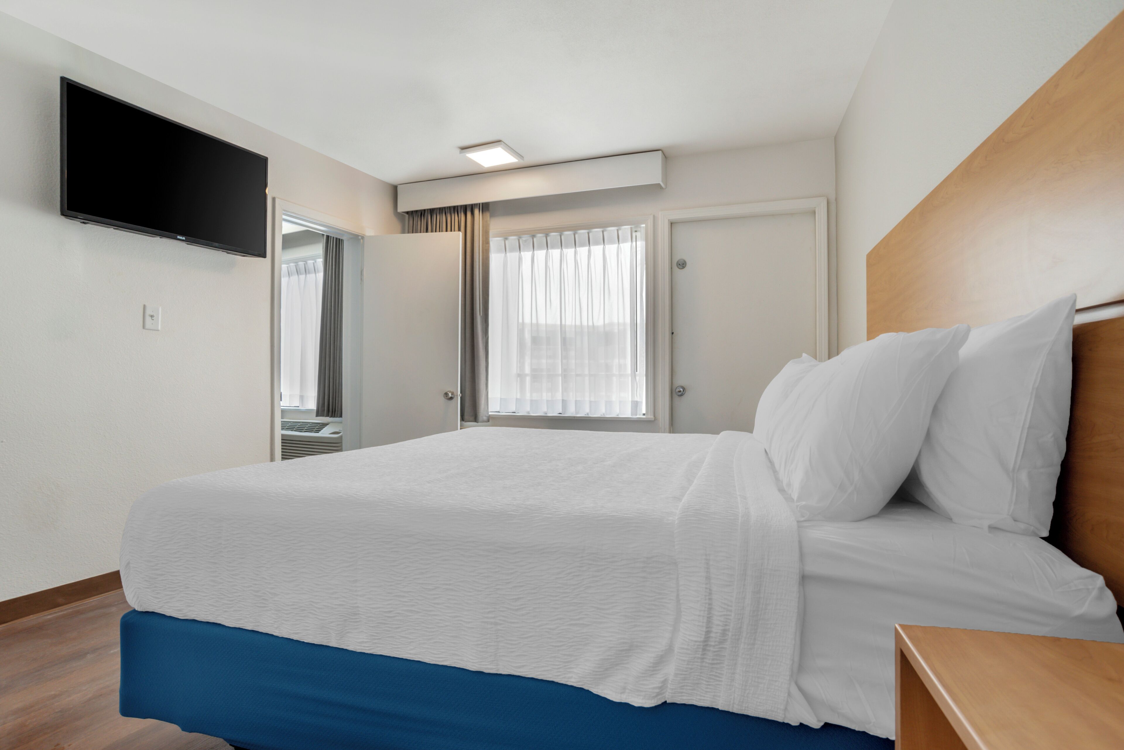 A hotel room with a bed, a TV mounted on the wall, a window with curtains, and a door.