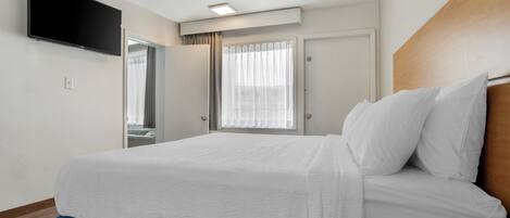 Una habitación de hotel con una cama, un televisor montado en la pared, una ventana con cortinas y una puerta.