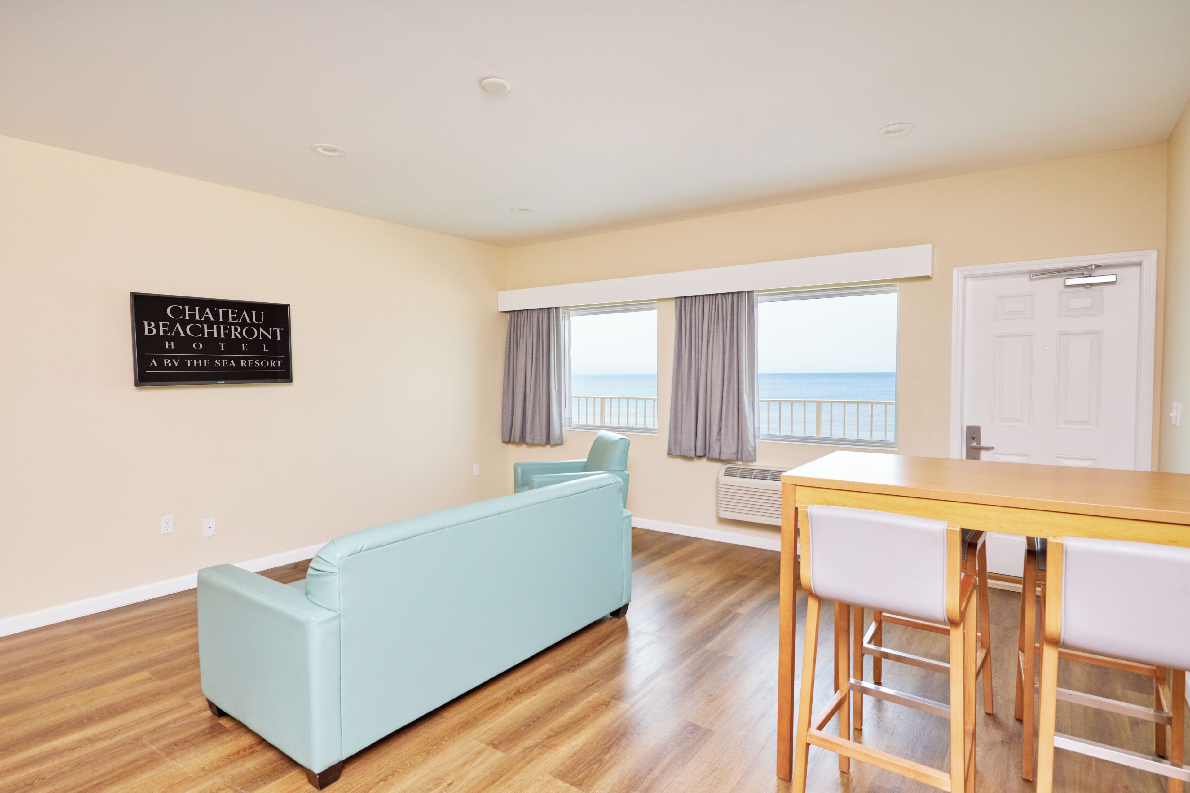 Un salon avec un canapé, une table à manger avec des tabourets de bar, une vue sur la mer et un panneau indiquant l’hôtel Chateau Beachfront.
