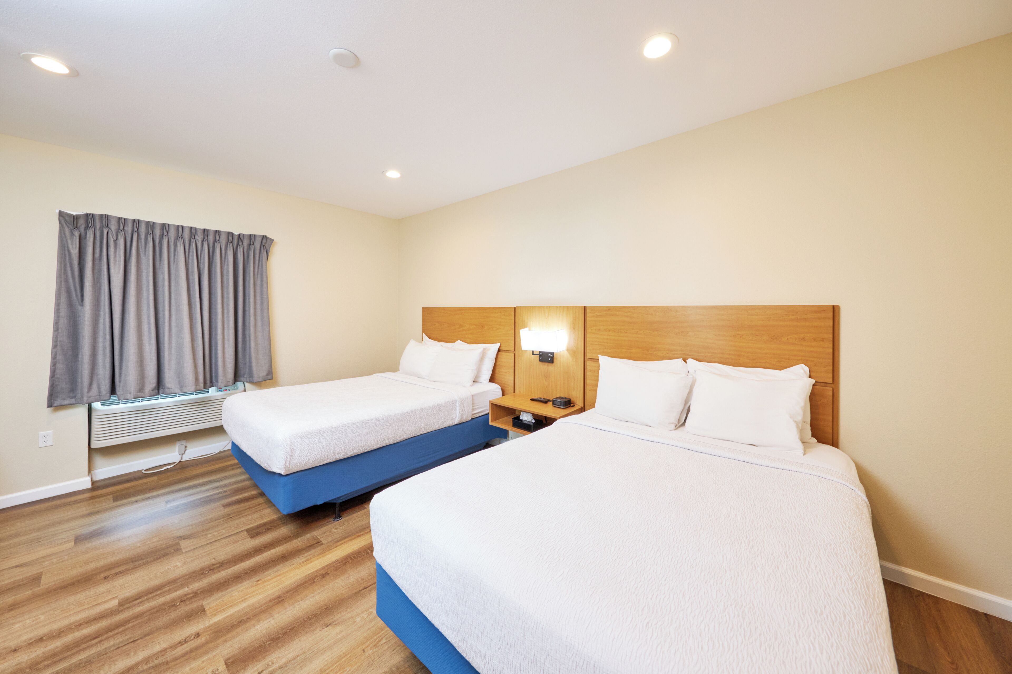 Une chambre d’hôtel avec deux lits, une tête de lit en bois, une table de chevet avec une lampe, une fenêtre avec des rideaux et un système de climatisation intégré.