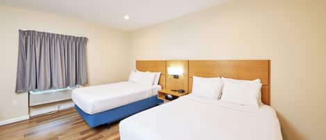 Habitación de hotel con dos camas, cabecero de madera, mesita de noche con lámpara, ventana con cortinas y aire acondicionado instalado.