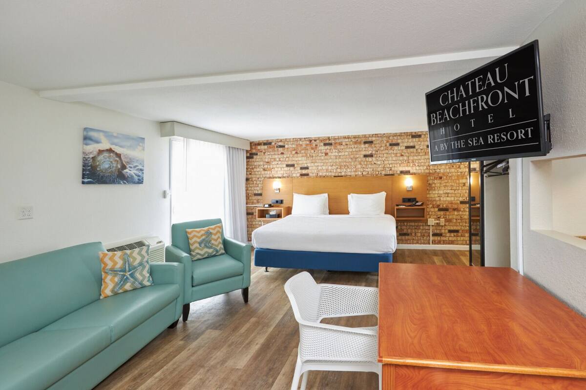 A hotel room with a bed, a teal sofa, a wooden table, and a brick-patterned wall.