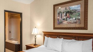 A hotel room with a bed, a nightstand with a lamp, a bathroom with a mirror, and a framed painting on the wall.