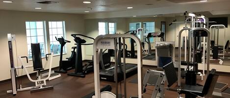 Uma academia com diversos equipamentos de exercício, incluindo esteiras e aparelhos de musculação, com amplas janelas que oferecem luz natural.