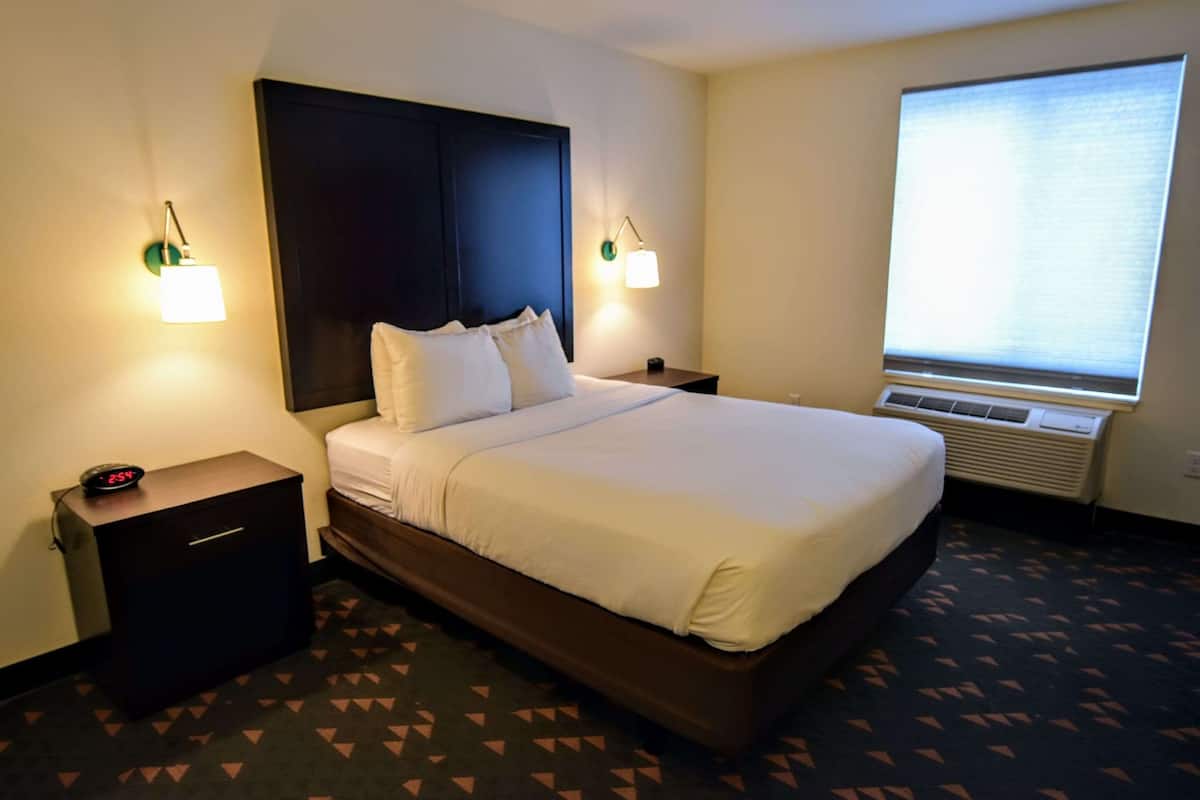 Une chambre d’hôtel avec un grand lit, deux tables de chevet, une petite table, une lampe fixée au mur et une fenêtre avec des stores.