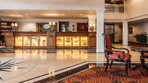 A spacious lobby with a reception desk, a chandelier, a patterned carpet, and a seating area with a chair and table.