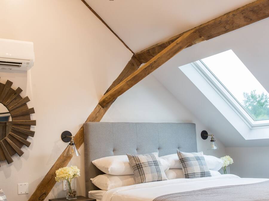 A bedroom with a vaulted ceiling, a skylight, a chandelier, a bed with multiple pillows, a bedside table with a lamp, and a decorative mirror.