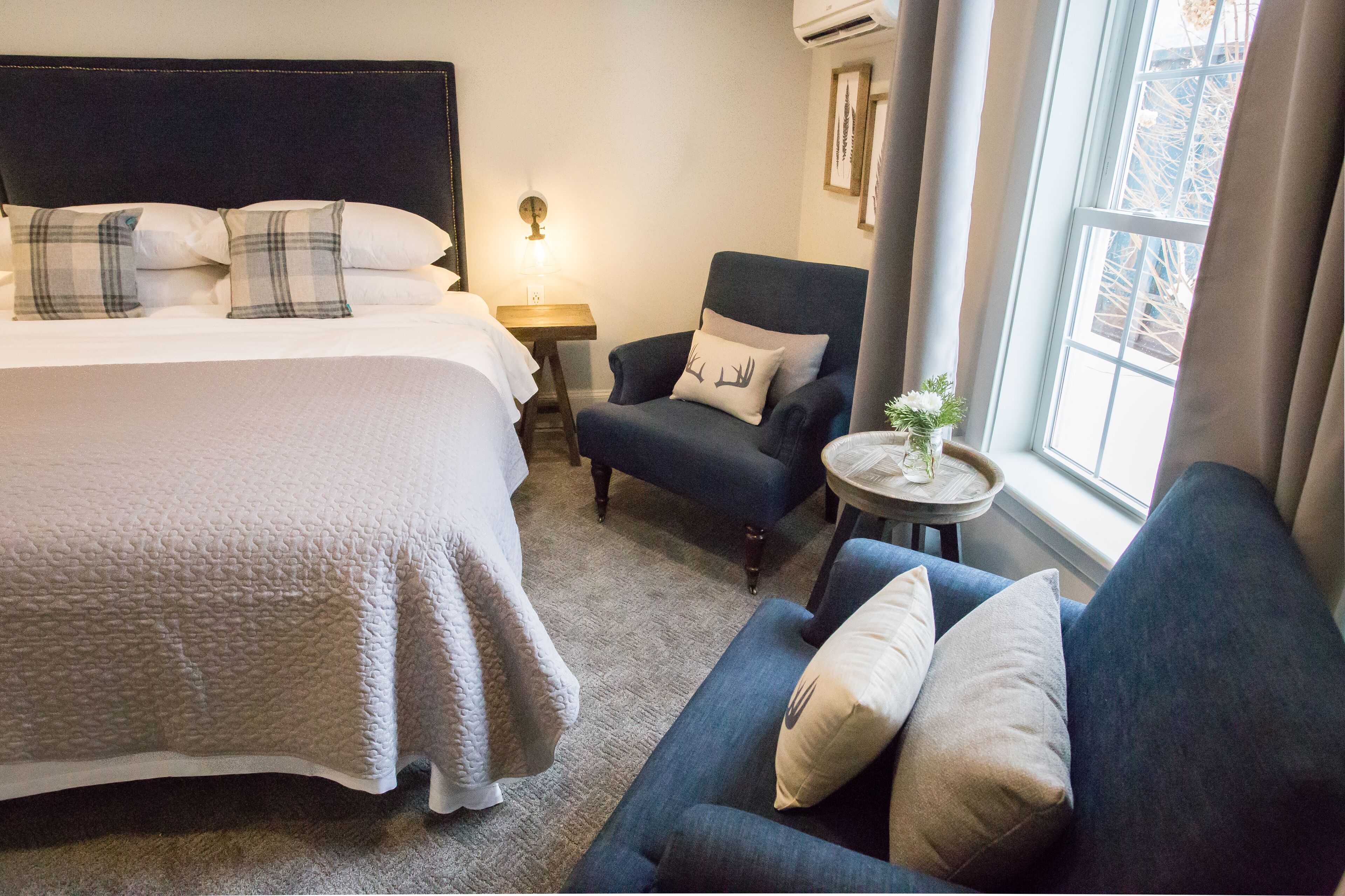 Una habitación de hotel con una cama, un sillón azul, una mesita de noche con una lámpara y un ventanal con cortinas.