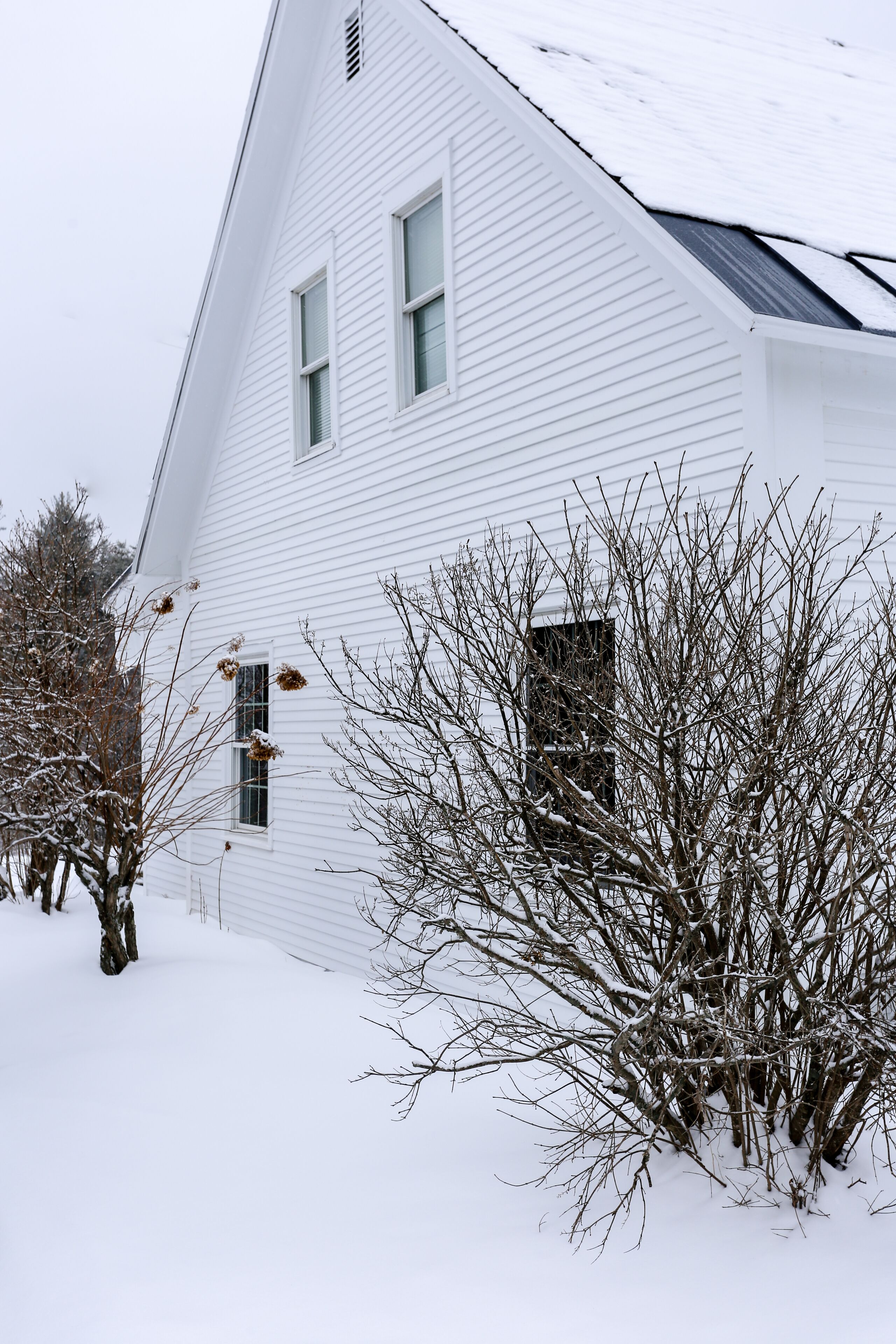 A white house with a steep roof, surrounded by snow-covered trees and bushes.