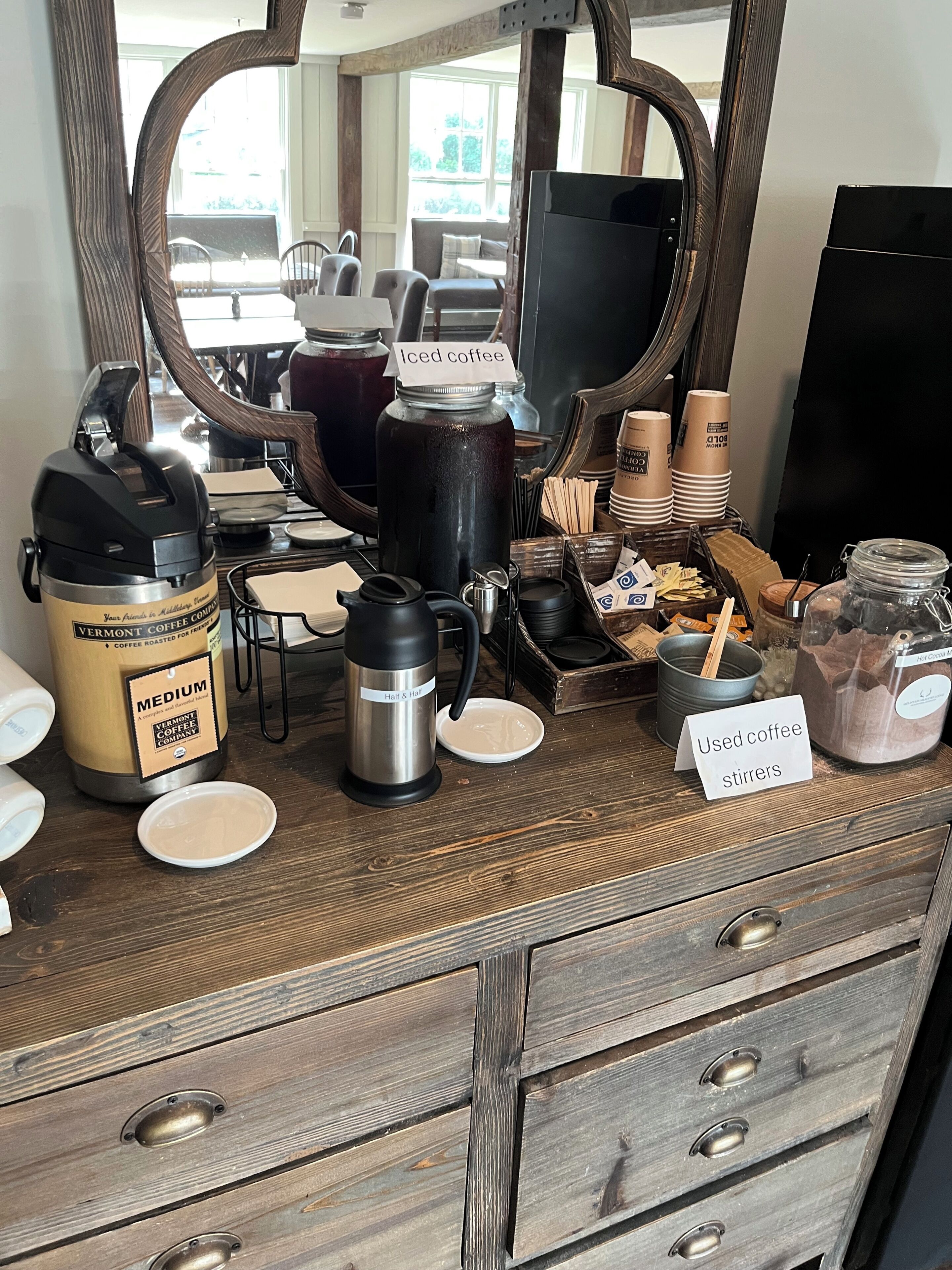 A coffee station with various brewing equipment and supplies, including a coffee maker, a thermos, and a jar of used coffee stirrers.